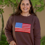 Woman wearing a brown sweatshirt with an American flag design outdoors.