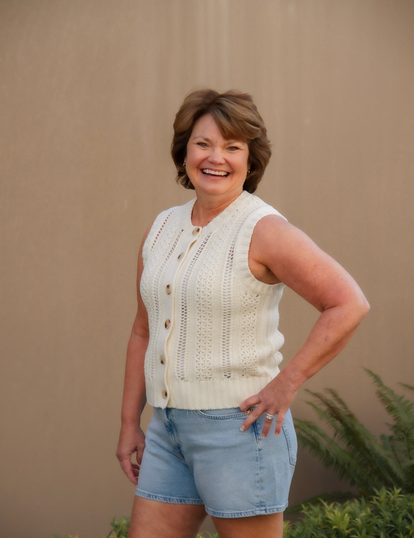Woman wearing a cream sleeveless top and light blue shorts against a beige wall.