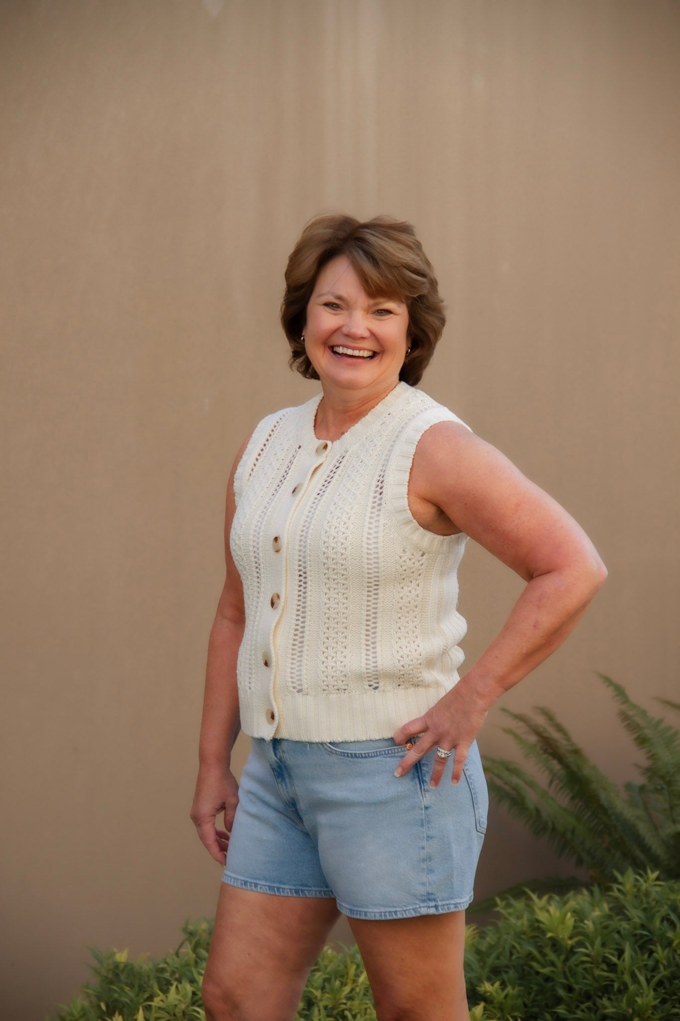 Woman wearing a cream sleeveless top and light blue shorts against a beige wall.