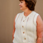 Woman wearing a cream sleeveless knitted top with buttons and light wash shorts against a beige wall.