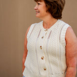 Woman wearing a cream sleeveless knitted top with buttons and light wash shorts against a beige wall.