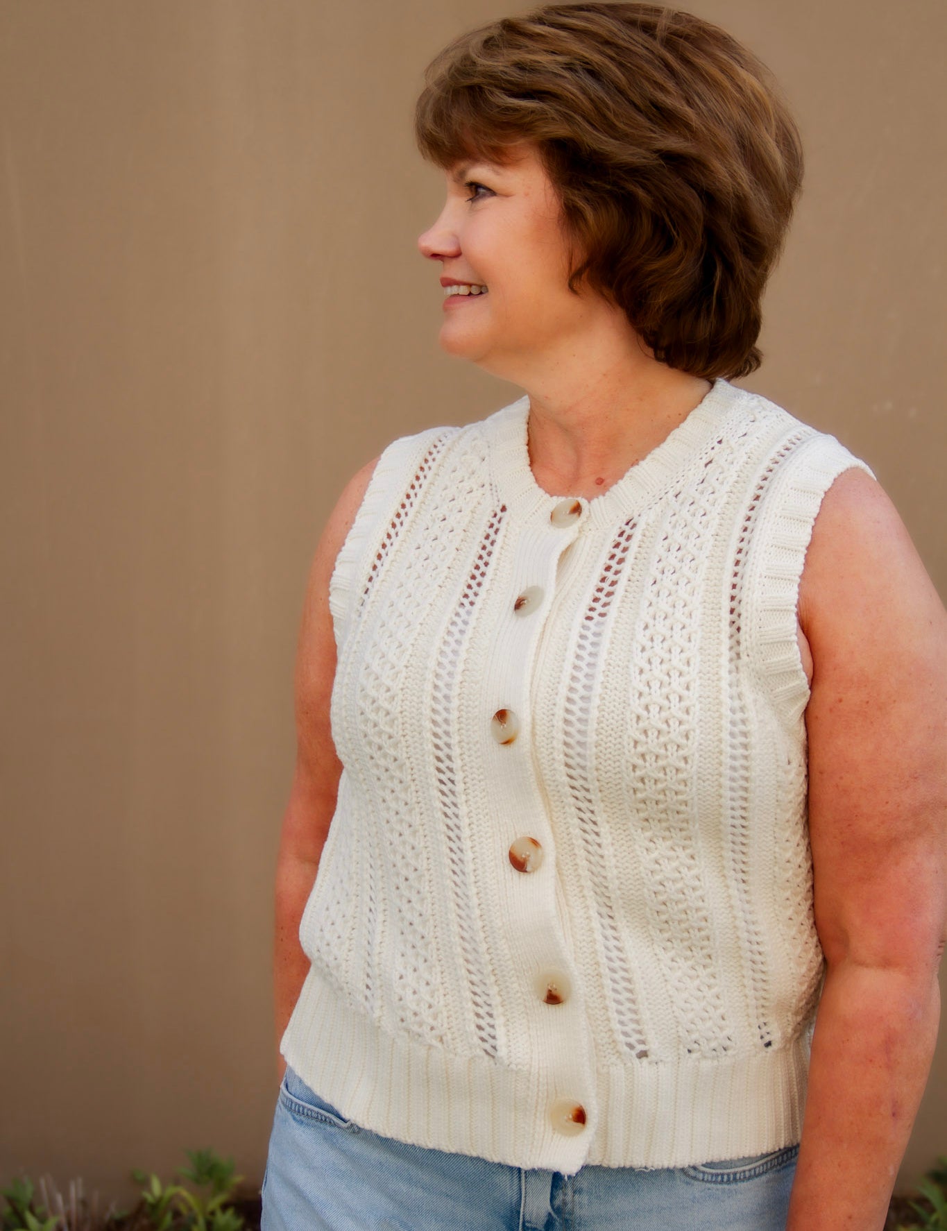 Woman wearing a cream sleeveless knitted top with buttons and light wash shorts against a beige wall.