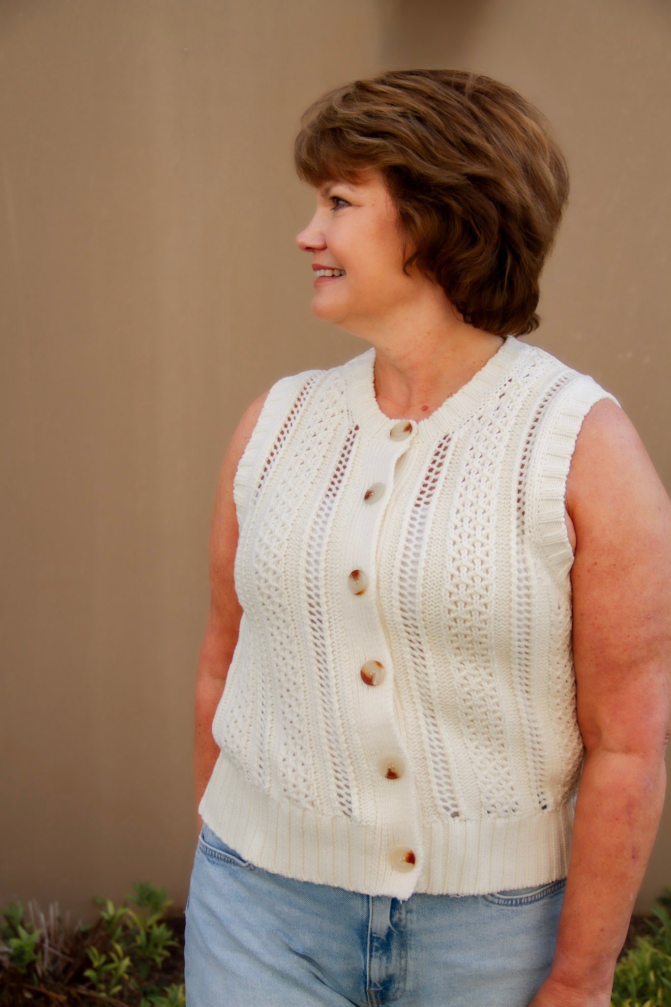 Woman wearing a cream sleeveless knitted top with buttons and light wash shorts against a beige wall.