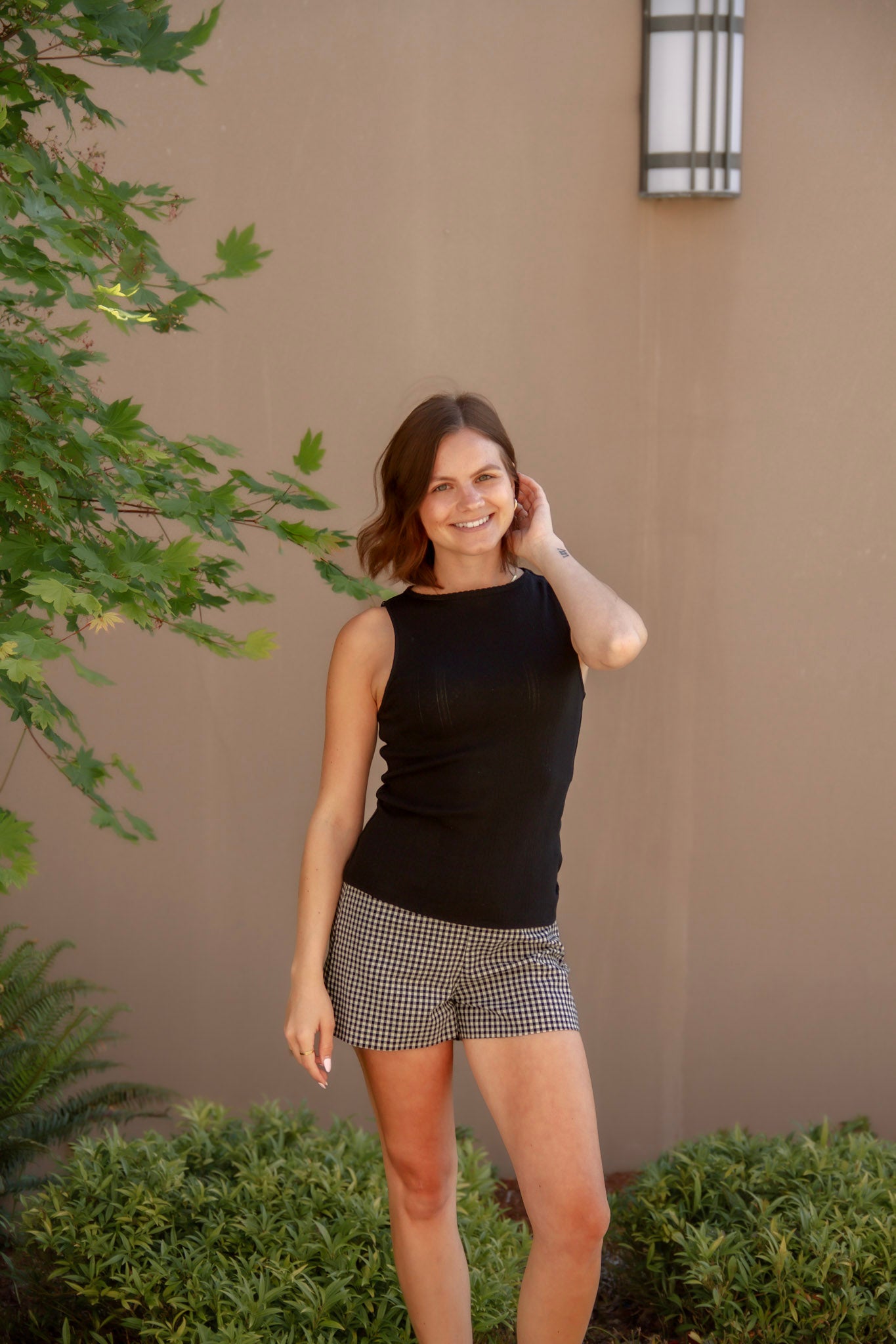 Woman in black sleeveless top and checkered shorts standing outdoors against a beige wall.