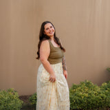 Woman wearing a plus size green top and yellow floral maxi skirt standing against a beige wall with greenery.