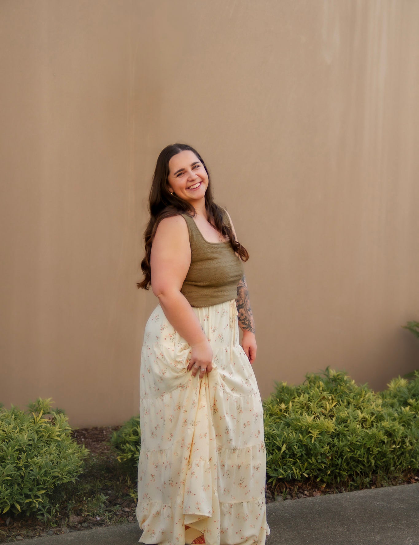 Woman wearing a plus size green top and yellow floral maxi skirt standing against a beige wall with greenery.