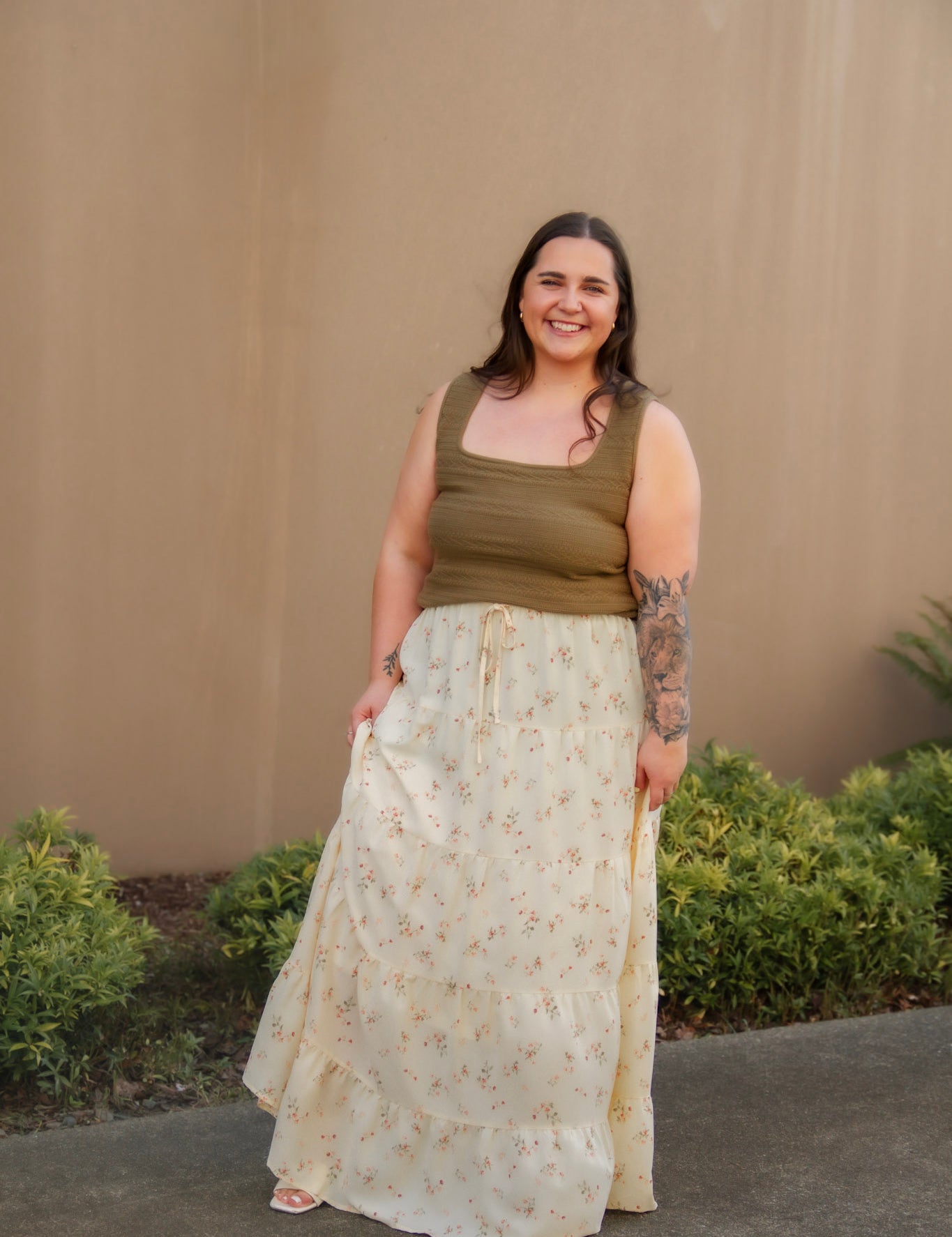 Woman wearing a green tank top and yellow floral skirt standing against a beige wall with plants in the background.