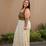 Woman wearing a green top and yellow floral skirt standing against a beige wall with greenery.