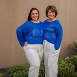 Two women wearing bright blue sweaters and white pants standing outdoors against a beige wall.