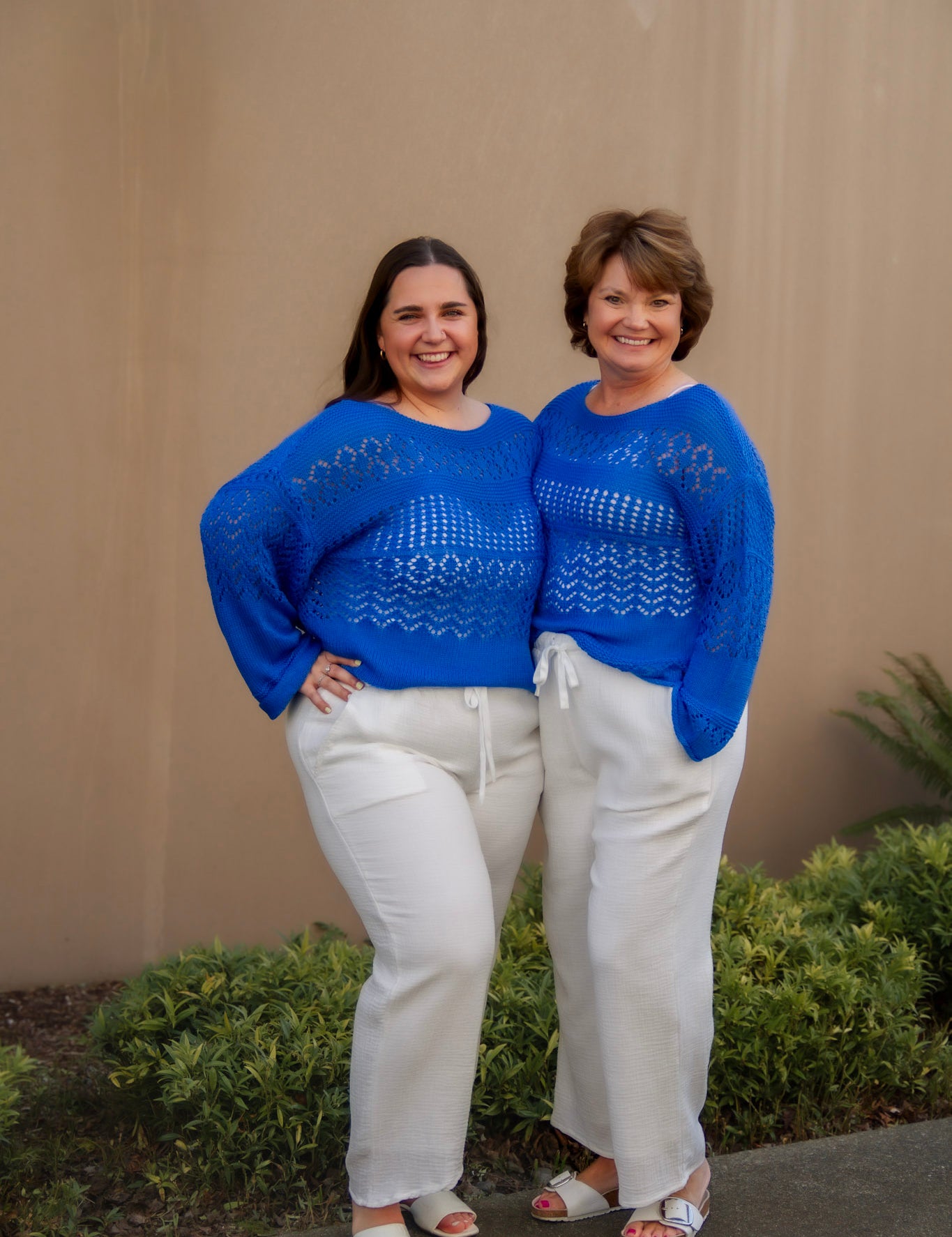 Two women wearing bright blue sweaters and white pants standing outdoors against a beige wall.