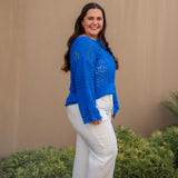 Woman wearing a blue top and white pants standing against a beige wall with greenery.