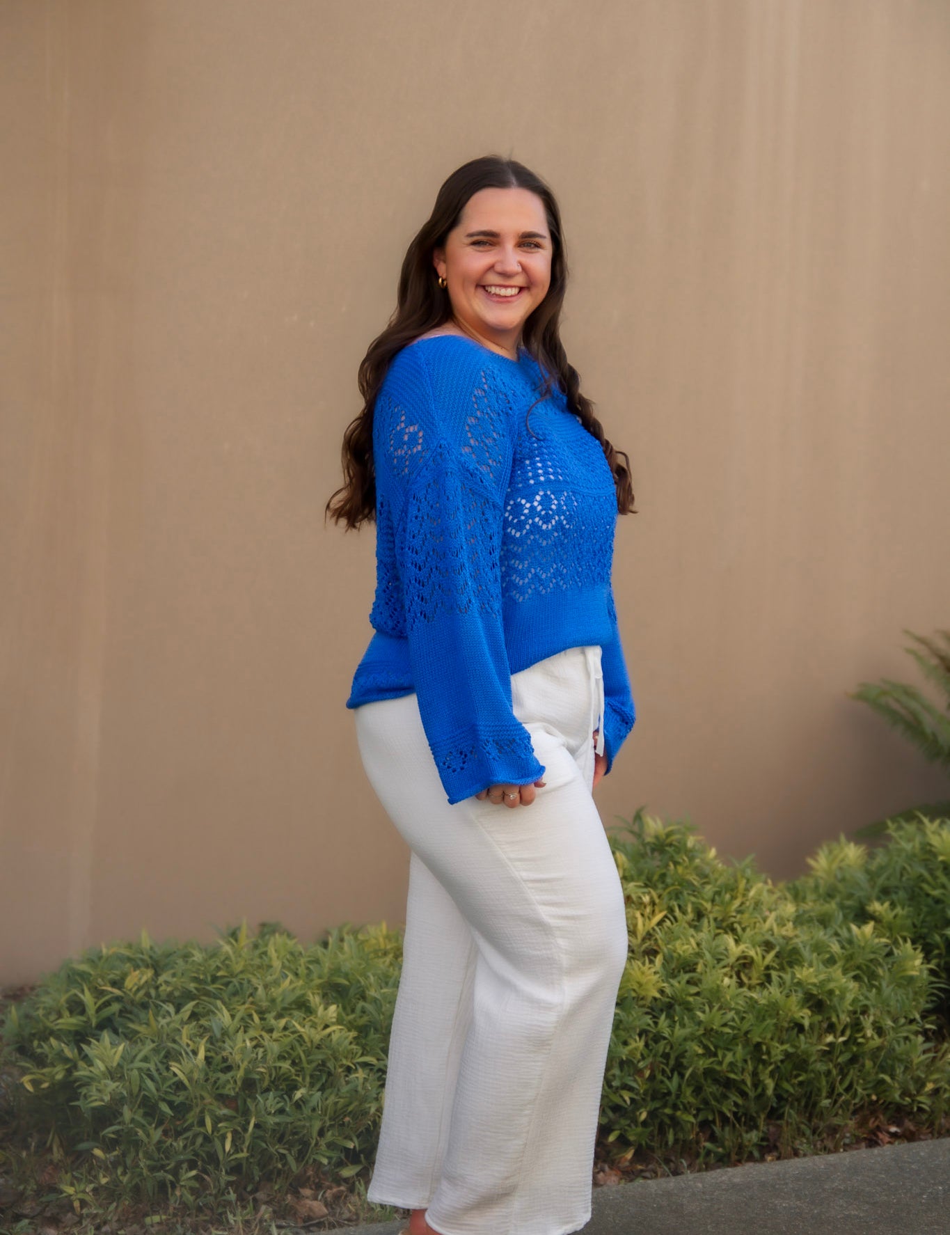 Woman wearing a blue top and white pants standing against a beige wall with greenery.