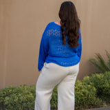 Person wearing a blue sweater and white pants standing against a beige wall with greenery.