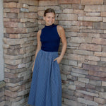 A woman stands in a stone-walled corner wearing a sleeveless navy blue mock-neck top and a blue-and-white vertically striped pleated midi skirt, paired with white slide sandals.