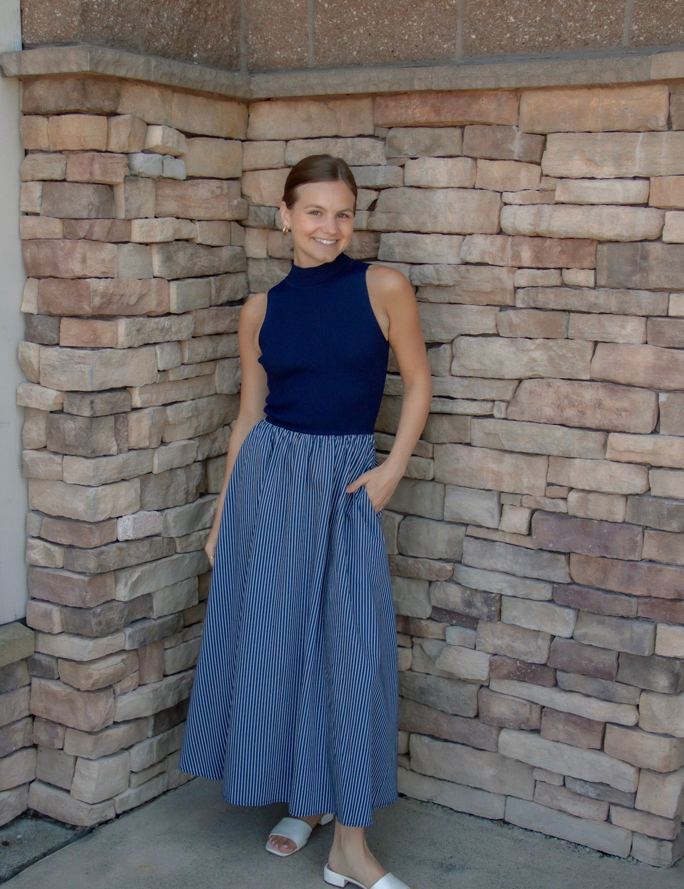 A woman stands in a stone-walled corner wearing a sleeveless navy blue mock-neck top and a blue-and-white vertically striped pleated midi skirt, paired with white slide sandals.