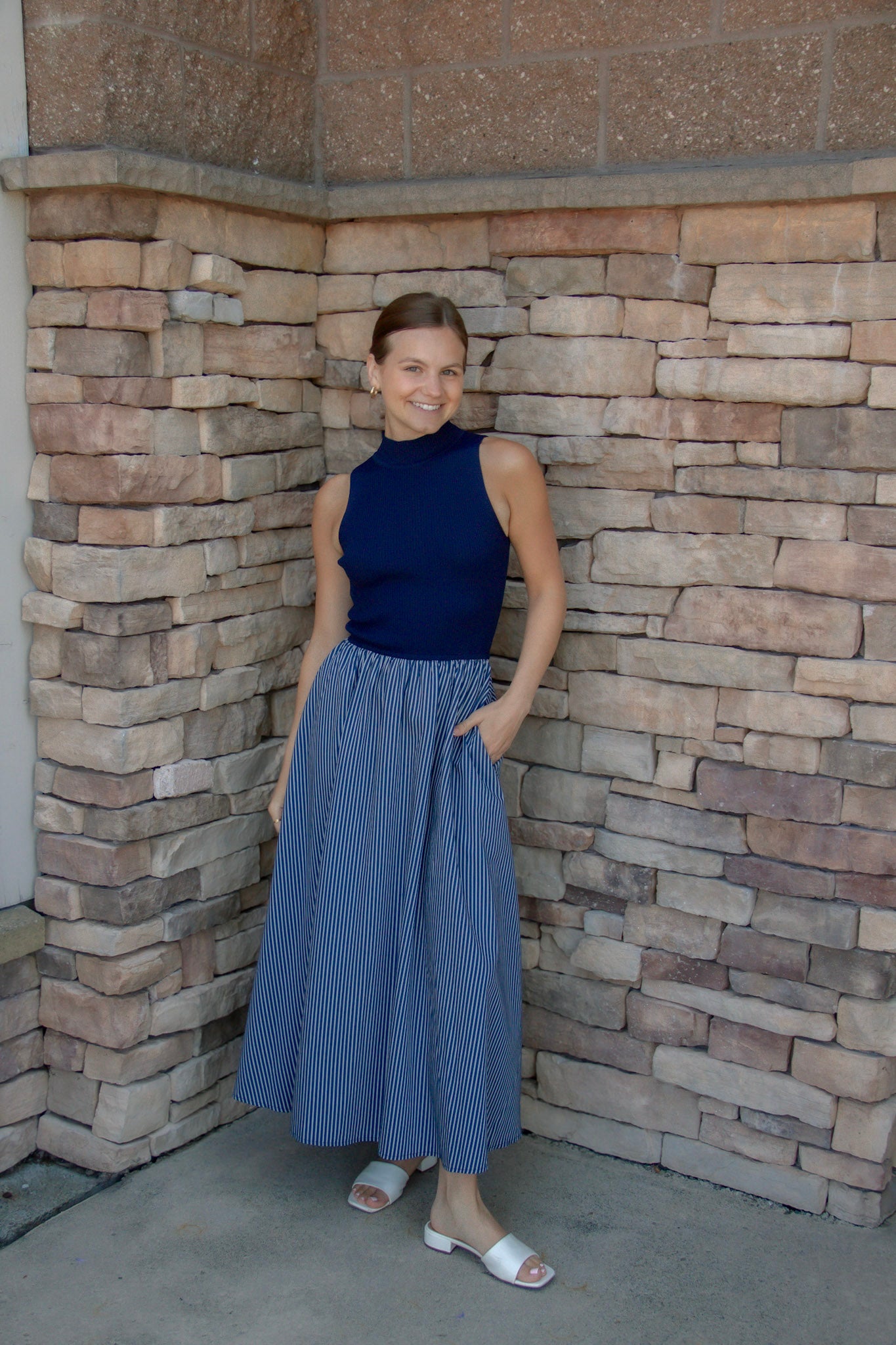 A woman stands in a stone-walled corner wearing a sleeveless navy blue mock-neck top and a blue-and-white vertically striped pleated midi skirt, paired with white slide sandals.