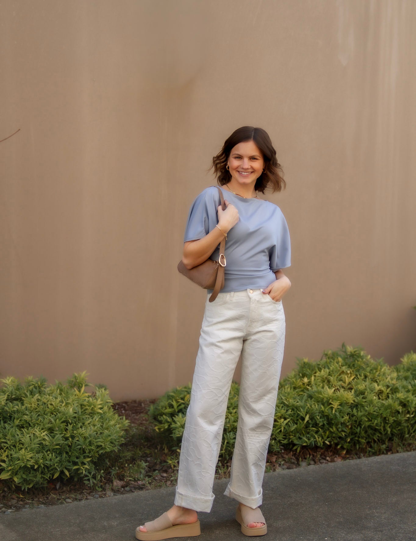 Woman in a blue shirt and white pants standing outdoors against a beige wall.