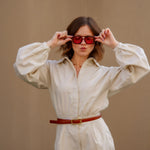 Woman wearing a beige shirt and shorts set with red sunglasses against a plain background