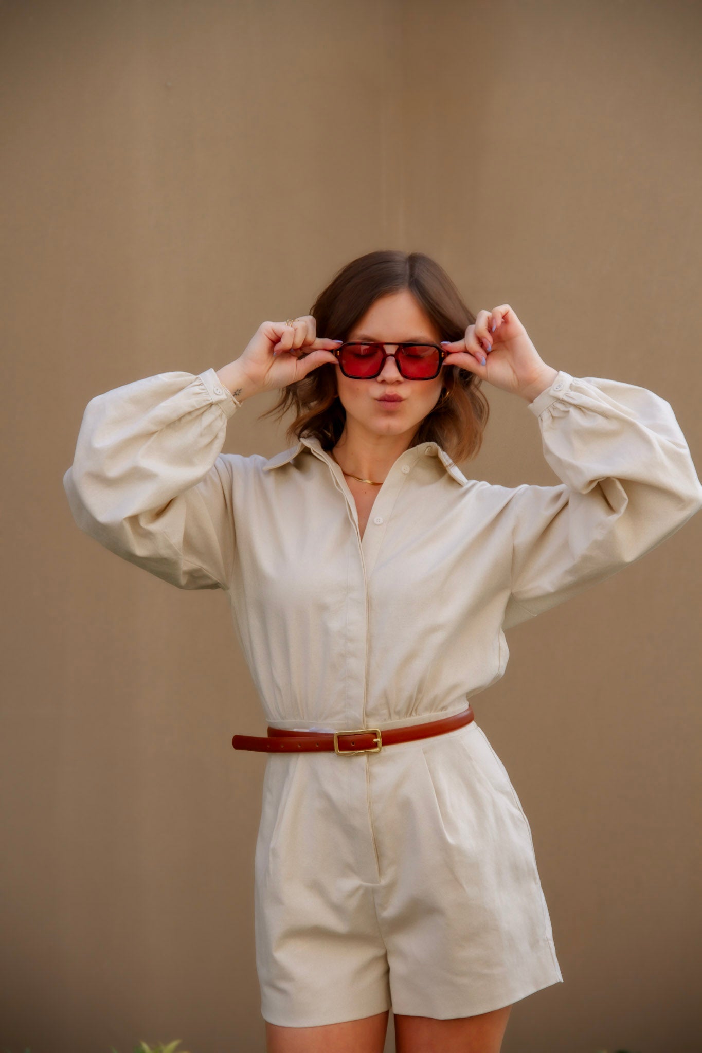 Woman wearing a beige shirt and shorts set with red sunglasses against a plain background