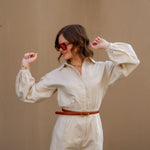 Woman wearing a tan romper and sunglasses with a brown belt against a beige wall.