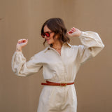 Woman wearing a tan romper and sunglasses with a brown belt against a beige wall.