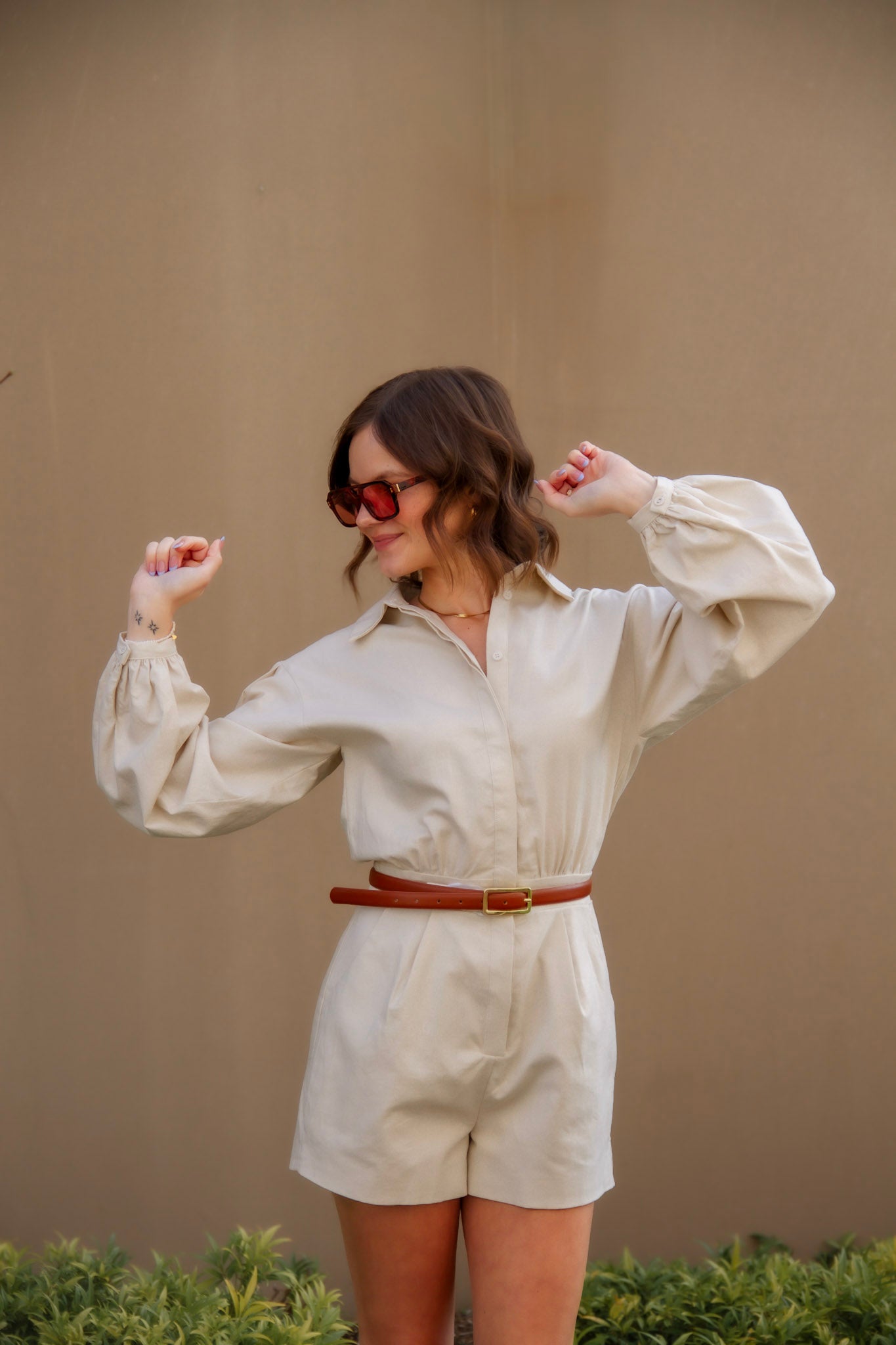 Woman wearing a tan romper and sunglasses with a brown belt against a beige wall.