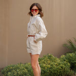 Woman in a tan romper outfit standing outdoors with a beige wall and greenery in the background