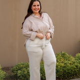 Woman in a beige striped long sleeve button down and white pants standing outdoors with a neutral wall and greenery in the background