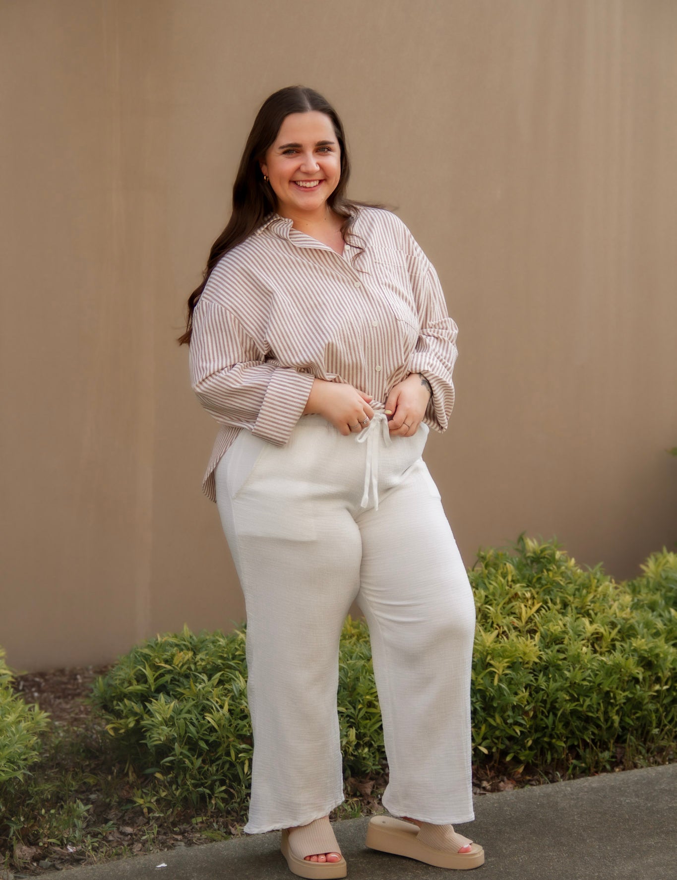 Woman in a beige striped long sleeve button down and white pants standing outdoors with a neutral wall and greenery in the background