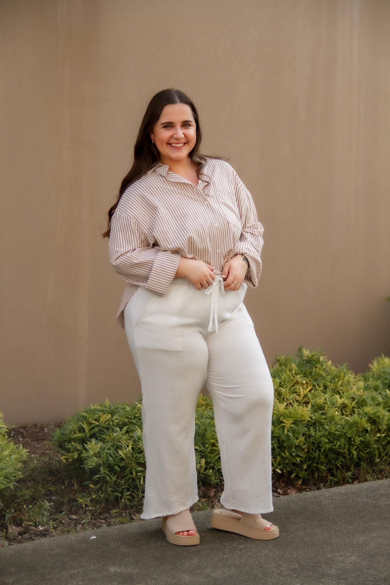 Woman in a beige striped long sleeve button down and white pants standing outdoors with a neutral wall and greenery in the background