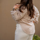 Woman wearing a beige striped shirt and white pants against a beige wall.