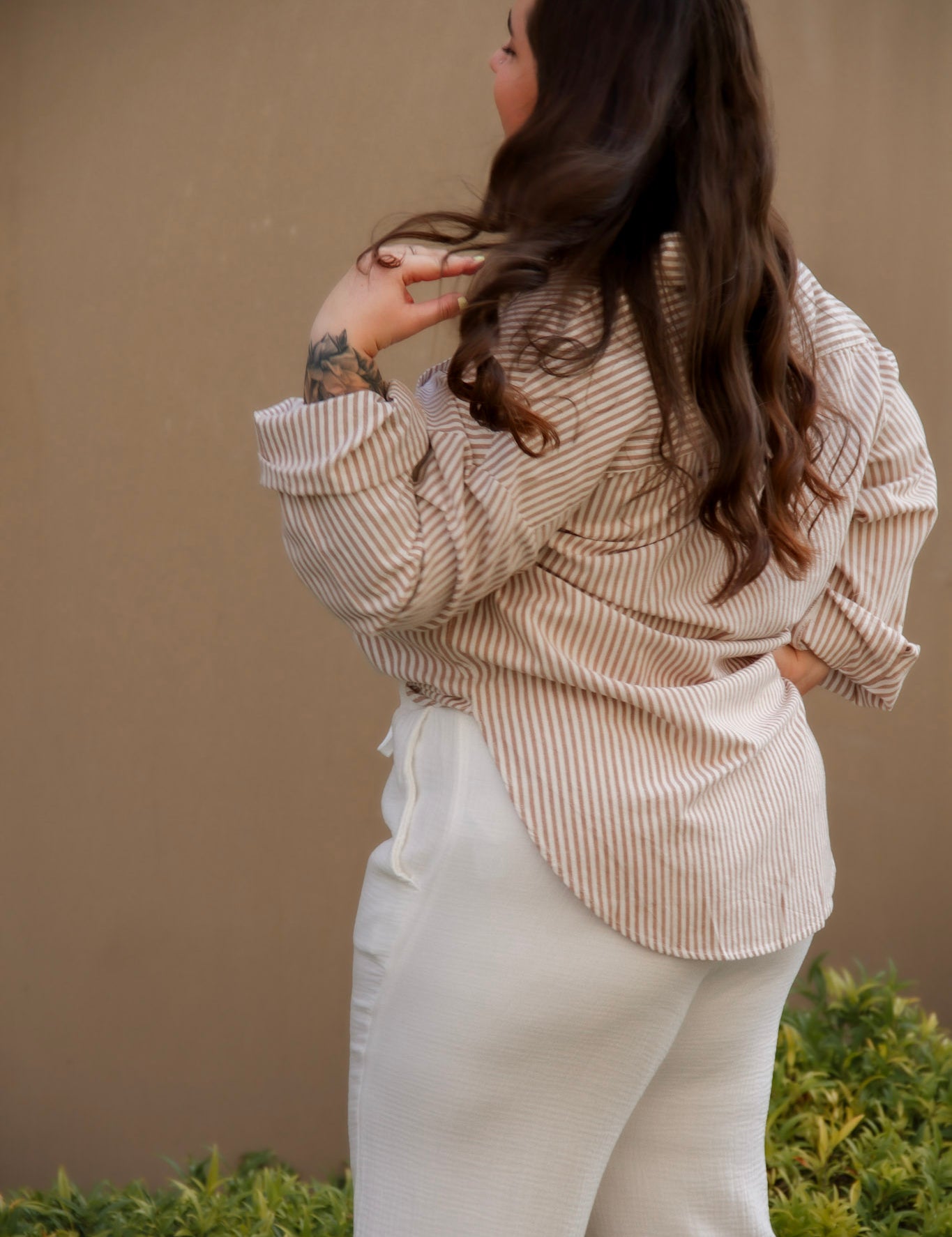 Woman wearing a beige striped shirt and white pants against a beige wall.