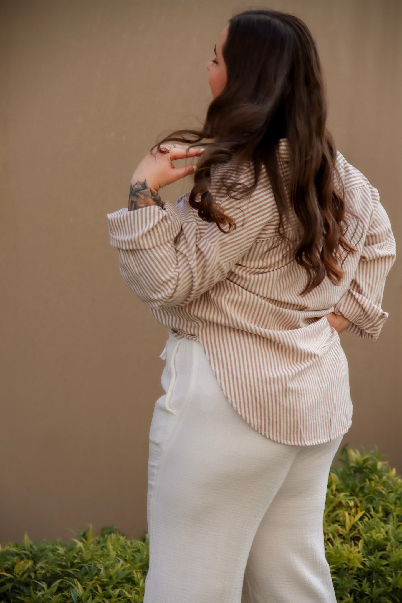 Woman wearing a beige striped shirt and white pants against a beige wall.