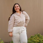 Woman wearing a striped shirt and white pants standing against a beige wall with greenery.