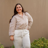 Woman wearing a striped shirt and white pants standing against a beige wall with greenery.