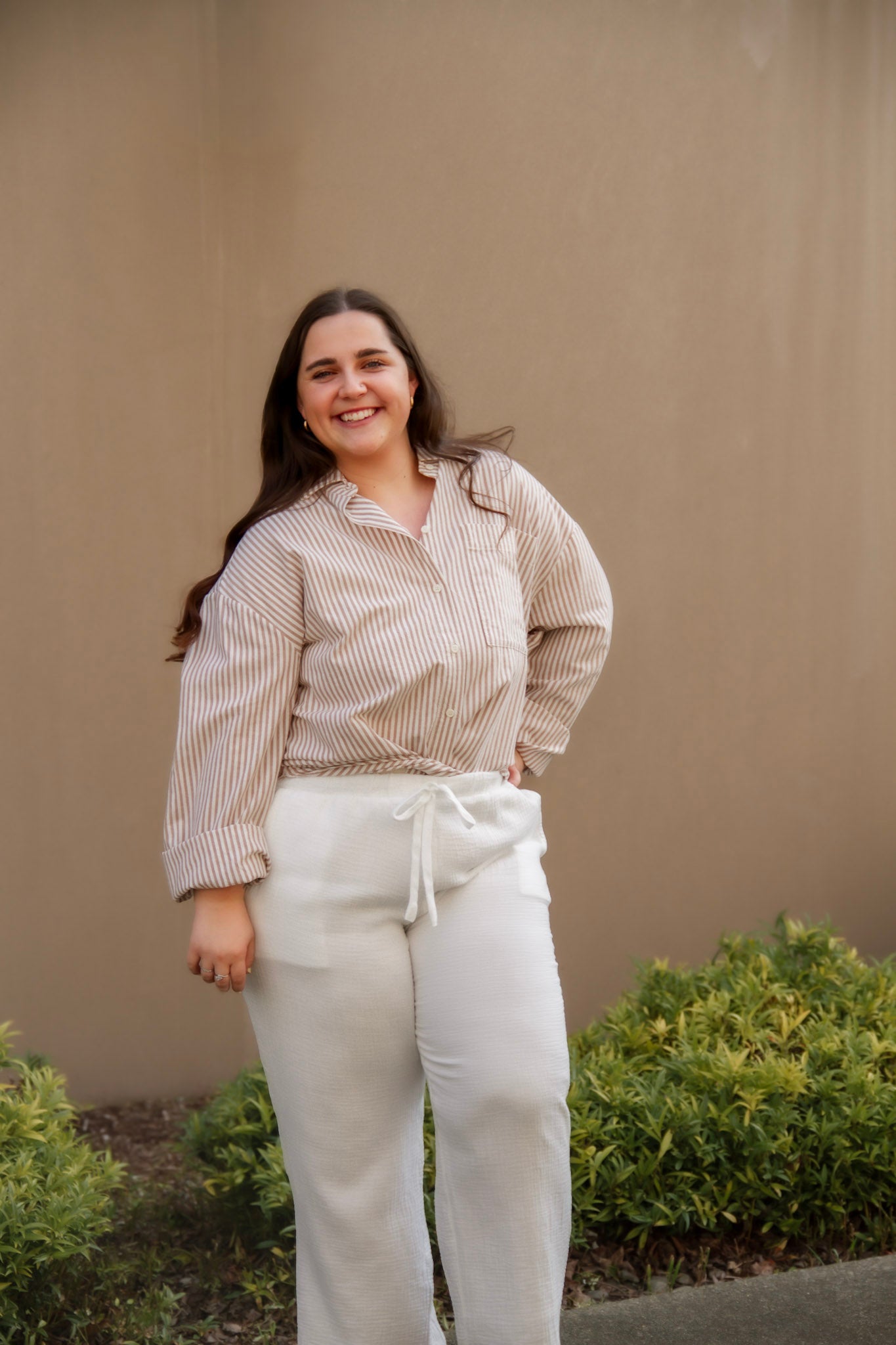 Woman wearing a striped shirt and white pants standing against a beige wall with greenery.