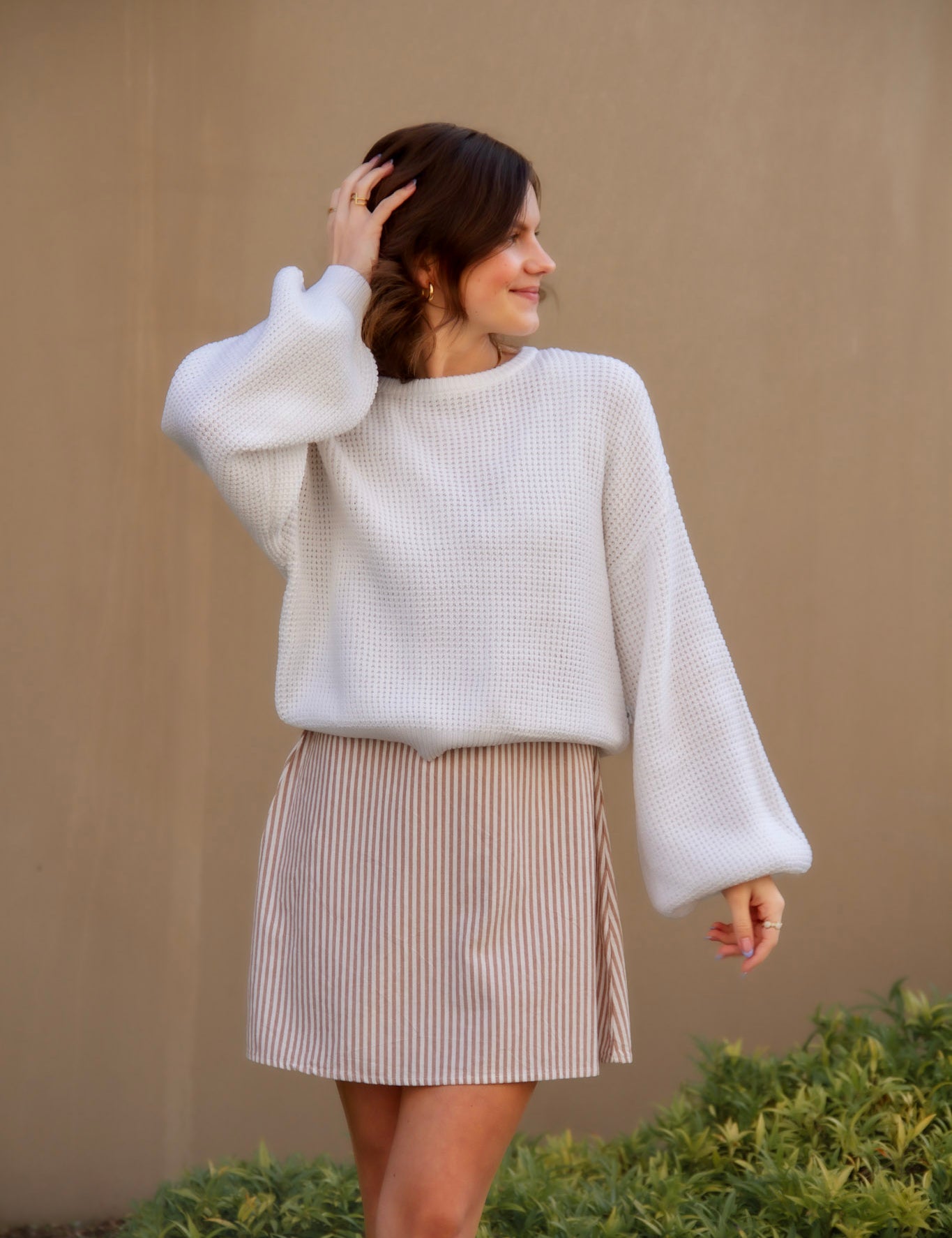 Woman wearing a white sweater and beige mini dress against a beige wall.