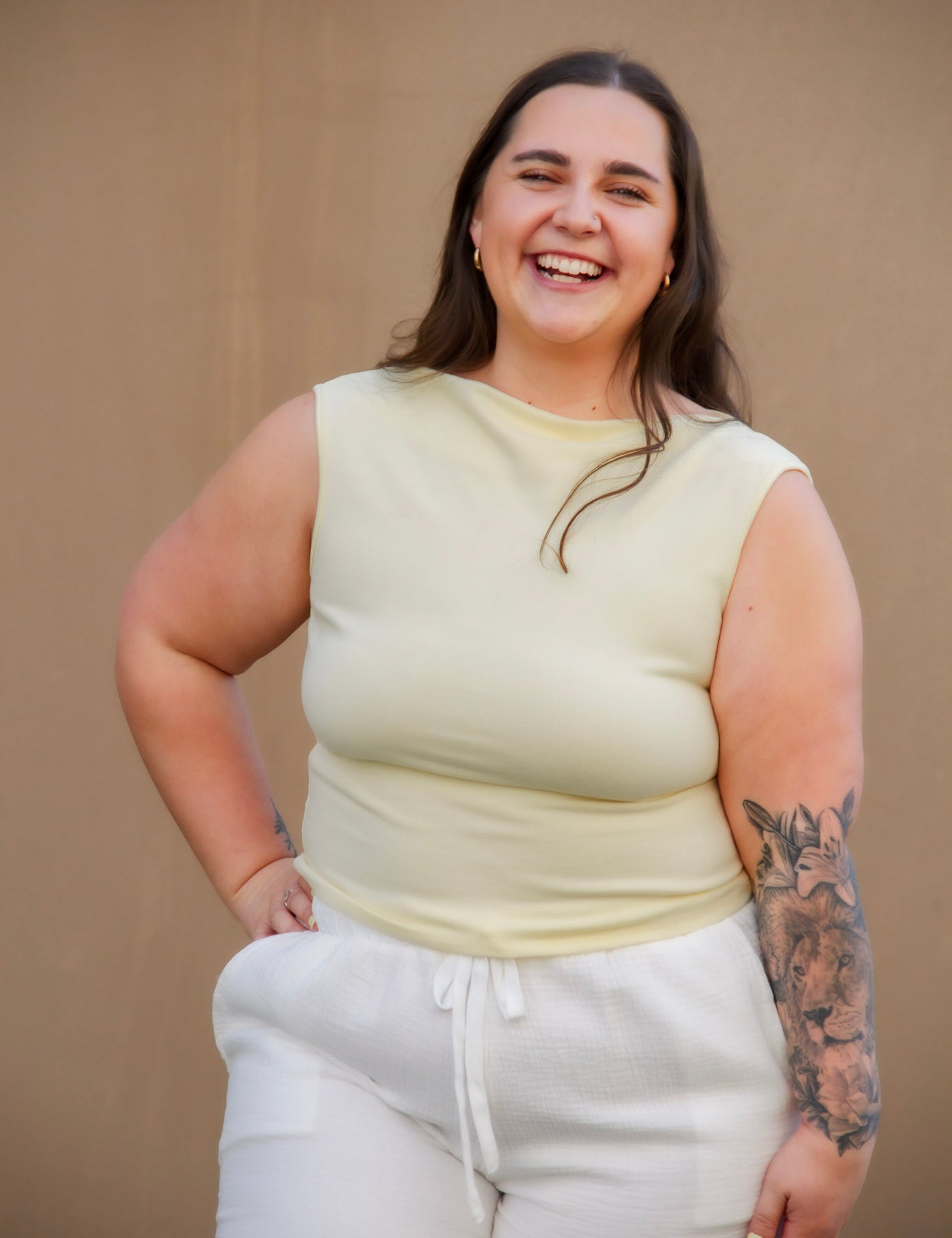 Woman wearing a light yellow sleeveless top and white pants against a beige background